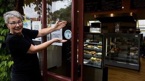 Photo shows smiling woman showing off the Refill NZ sticker in the window of her cafe.