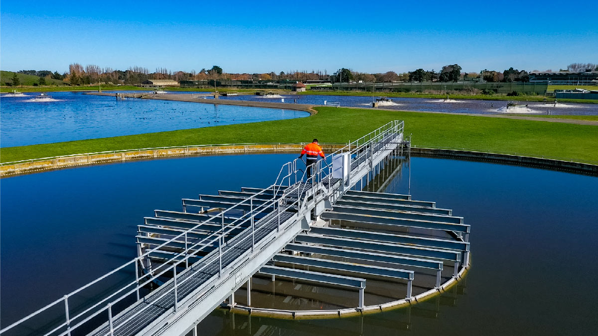 Image shows a man in high-res vest walking on a bridge over the waste water treatment plant