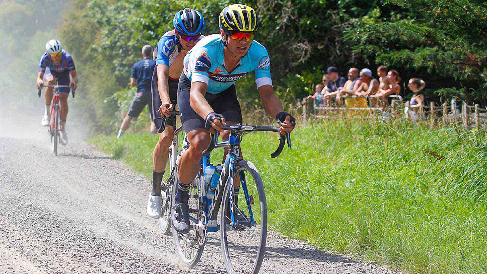 Photo shows people watching as cyclists race bikes along a gravel road, stirring up clouds of dust.
