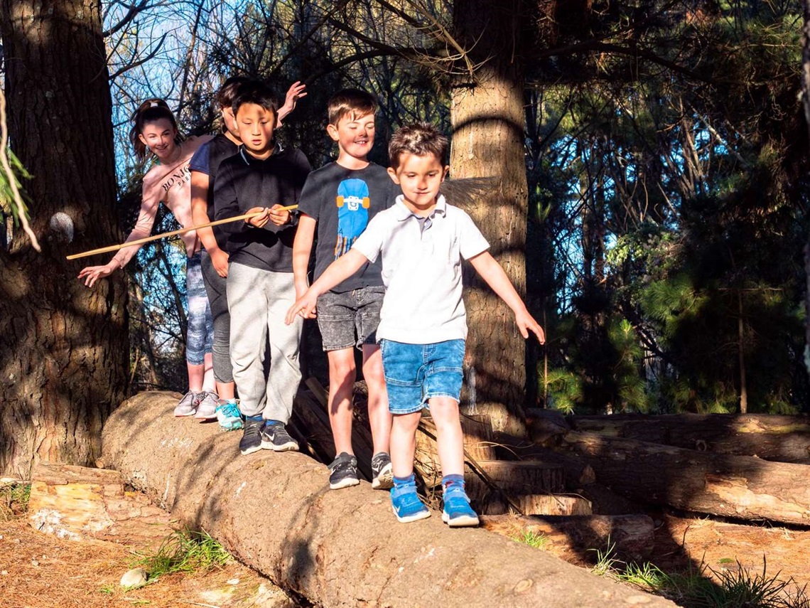 Photo shows five kids with excellent balance walking in single file along a large log in the bush.