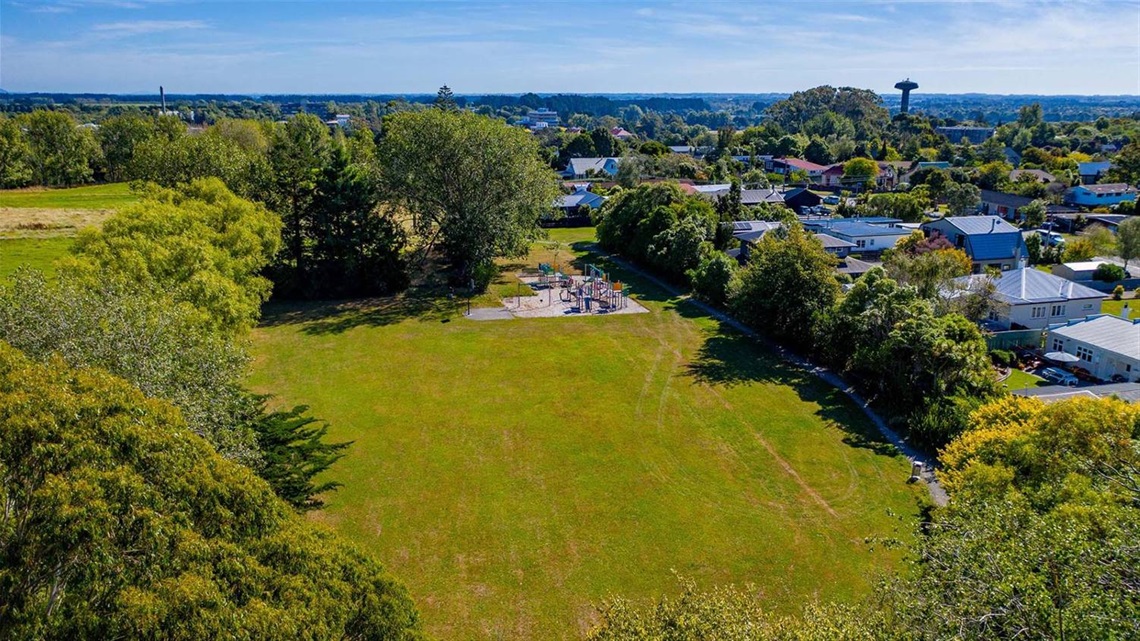 Photo shows an aerial view of a small open grassed space surrounded by large leafy trees and a playground at the far end.