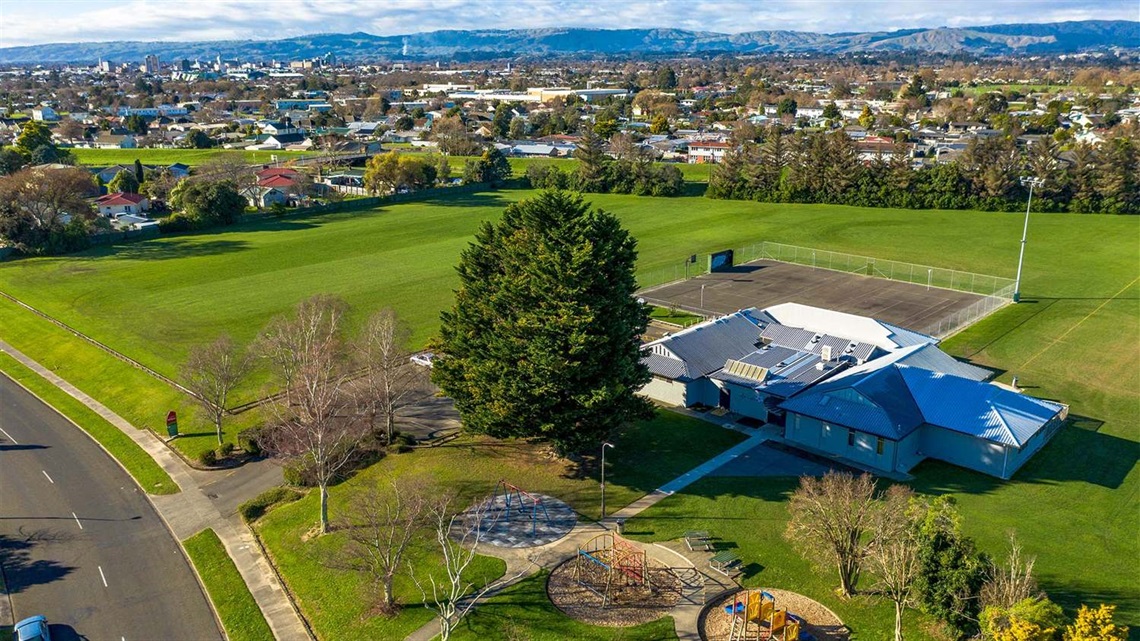 Photo shows large grassed area edged with trees. There is a small playground, a basketball court and a community centre.