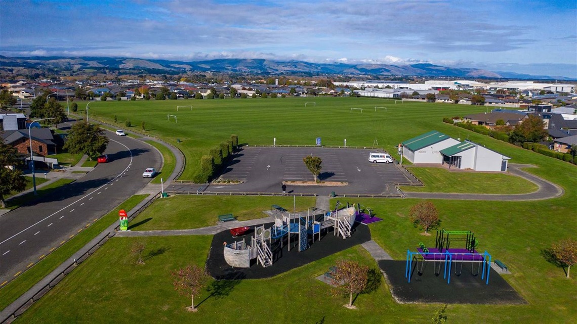 Photo shows a flat grassed area with a sportsfield in the background and a cool playground featuring a Viking ship.