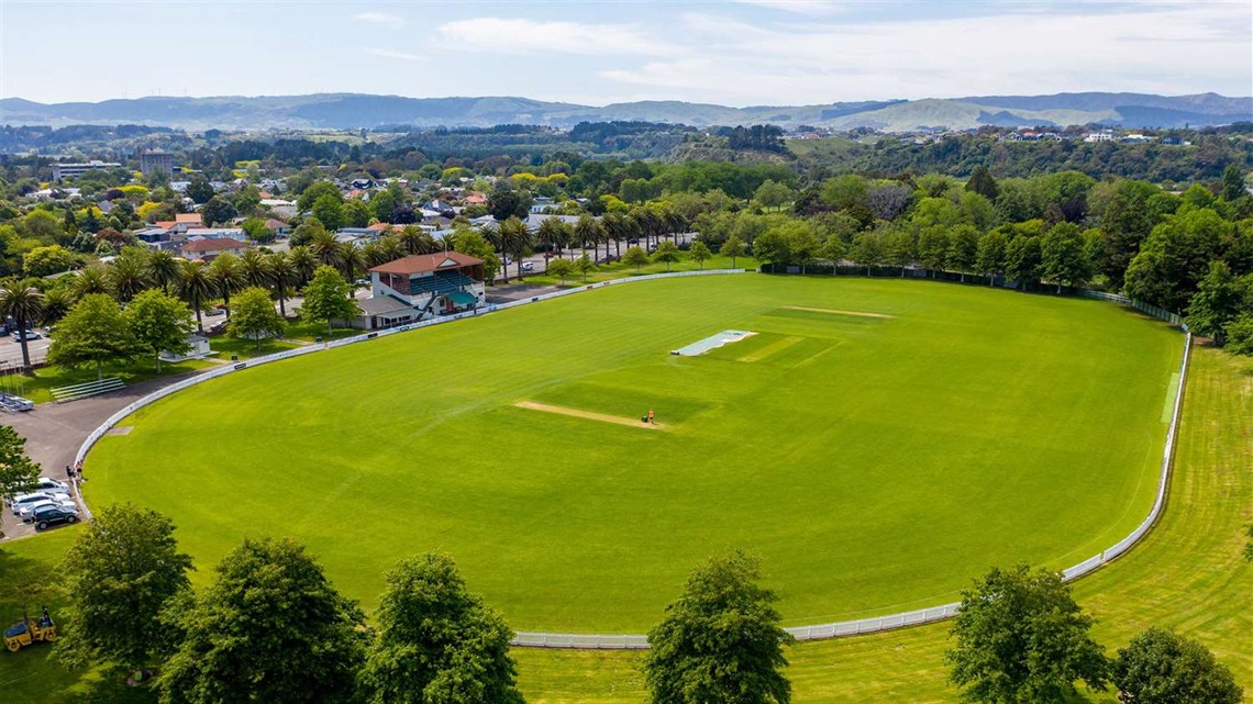 Photo shows cricket oval with pavilion in the background, surrounded by established leafy trees and palms.