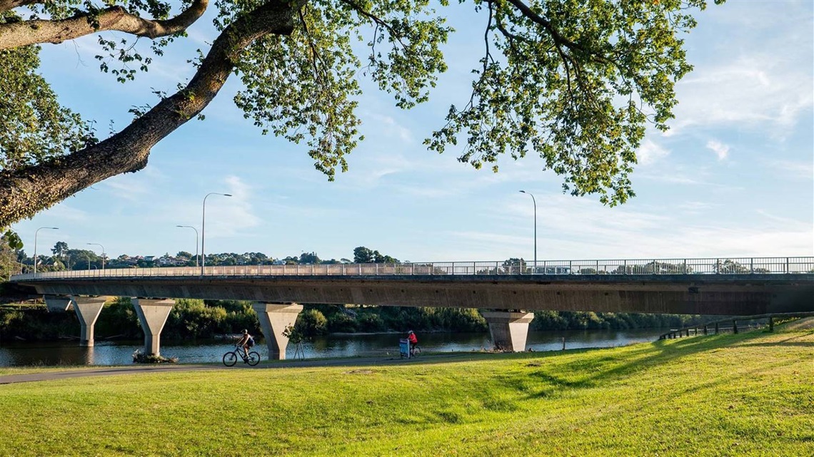 Photo shows a flat grassed area flanked by the stopbank on one side and the riverside walkway on the other. Fitzherbert Bridge is in the background.