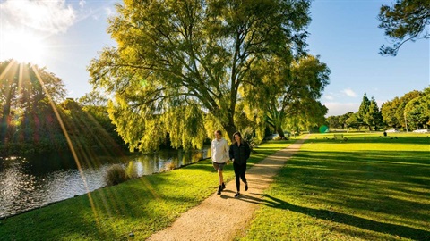 Couple walking along limestone pathway with lagoon on one side and grassy reserve full of trees on the other.