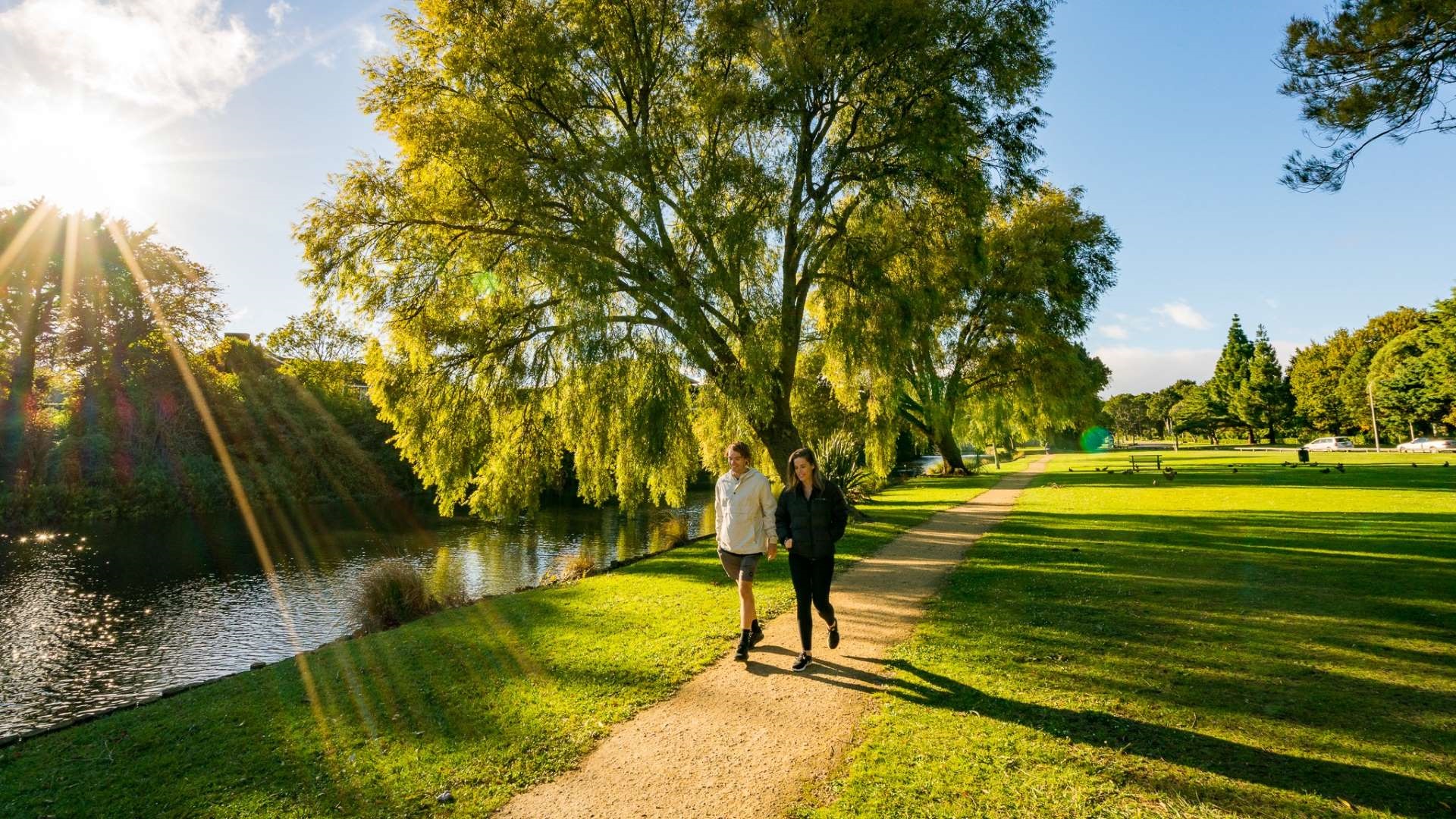 Photo shows couple walking along limestone pathway with lagoon on one side and grassy reserve full of trees on the other.