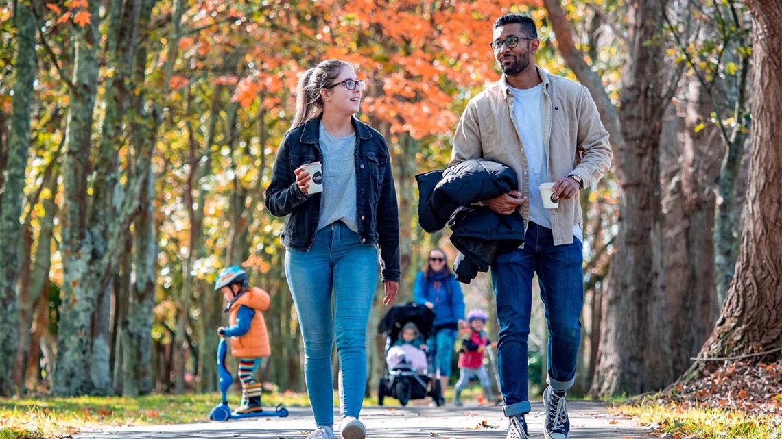 Photo shows people enjoying the tree-lined pathway in autumn: a young couple with takeaway coffee in hand, a woman pushing a baby in a stroller and two toddlers on kick-scooters.
