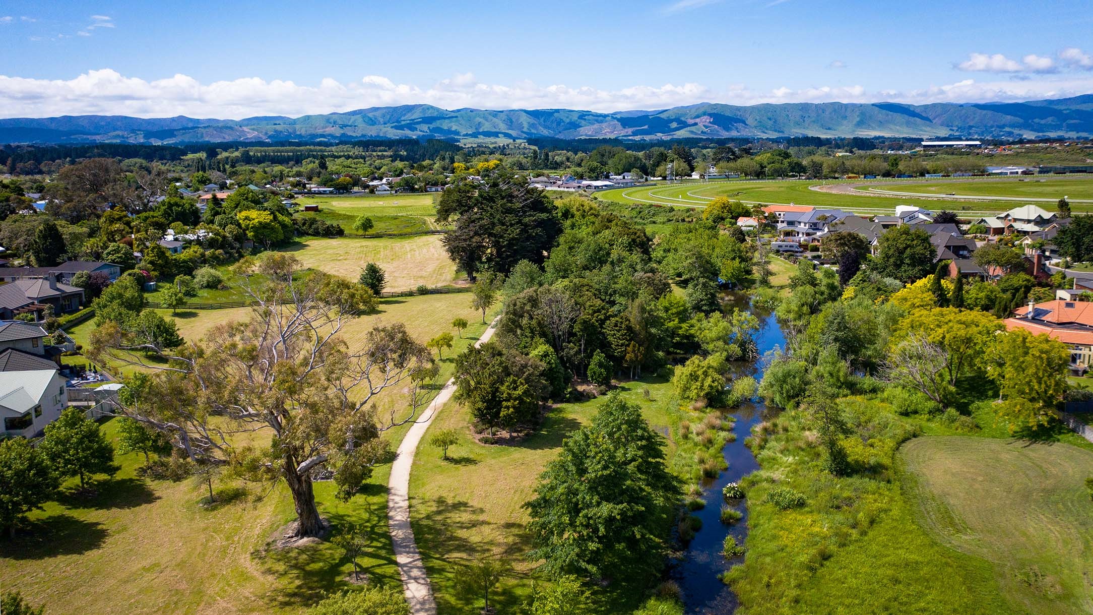 Photo shows aerial view of reserve with a stream and a limestone walkway running through it and a large flat grassed area that might be sportsfields in the background.