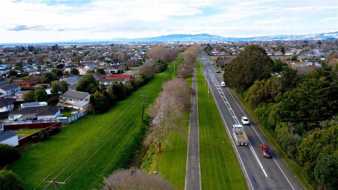 Photo shows flat grassed area and off-road shared pathway running adjacent to the state highway, bordered by homes on the left and the road on the right..