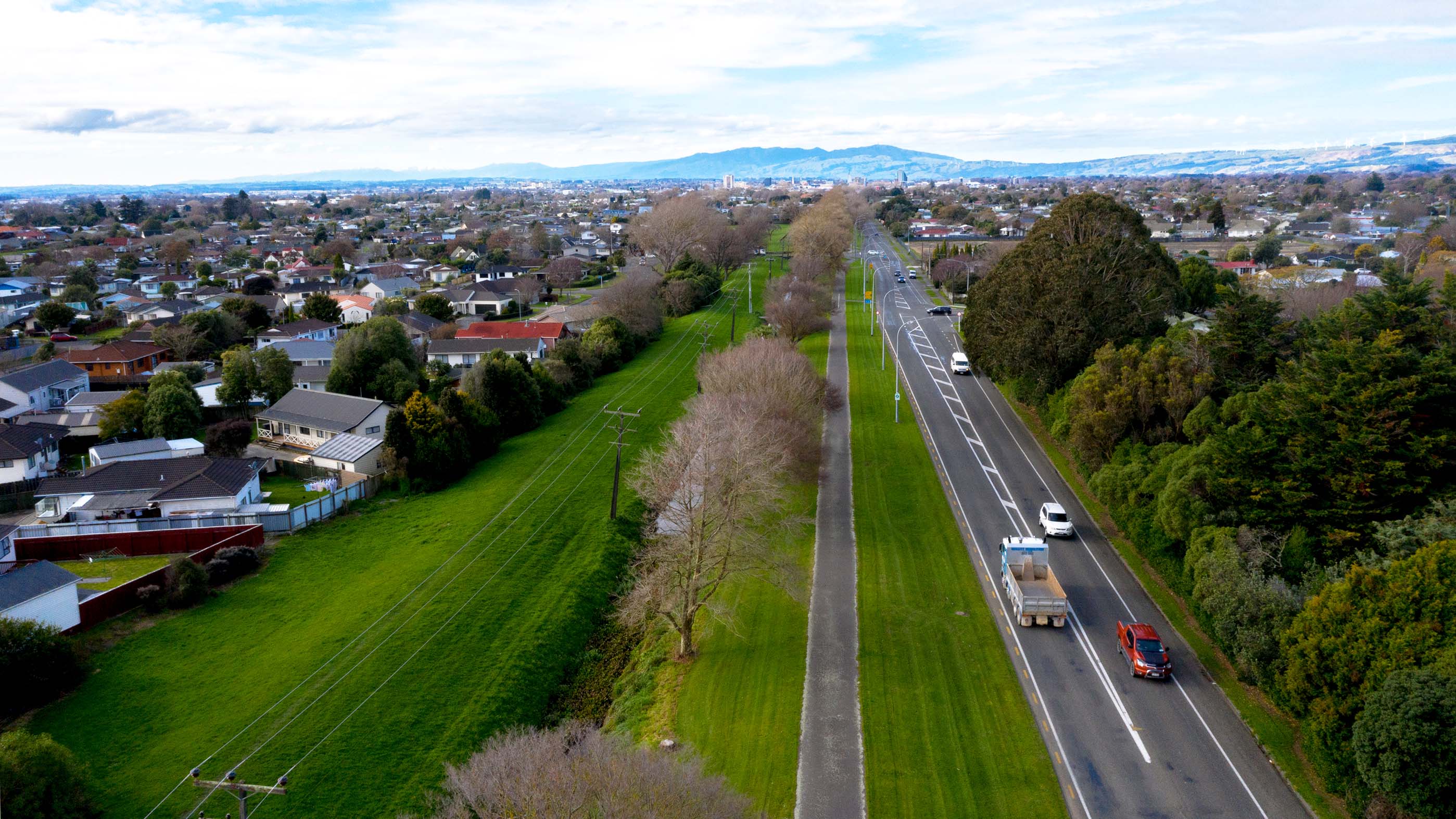 Flat grassed area and off-road shared pathway running adjacent to the state highway, bordered by homes on the left and the road on the right..