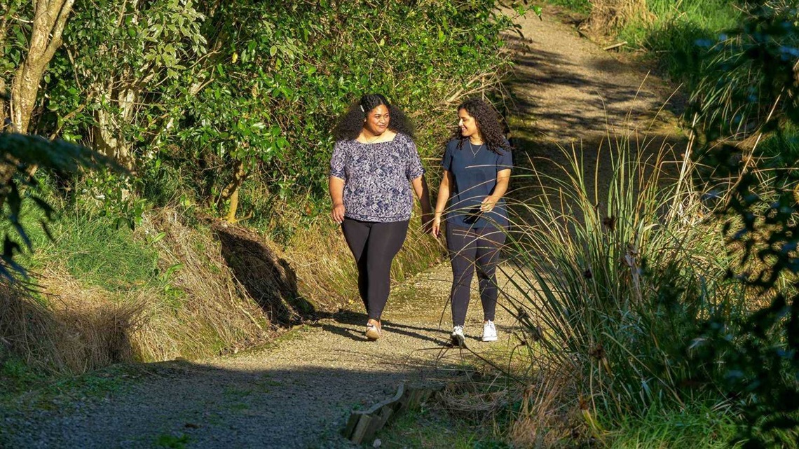 Photo shows two young women talking as they walk along a gravel path which is rising steeply out of a bushy valley.