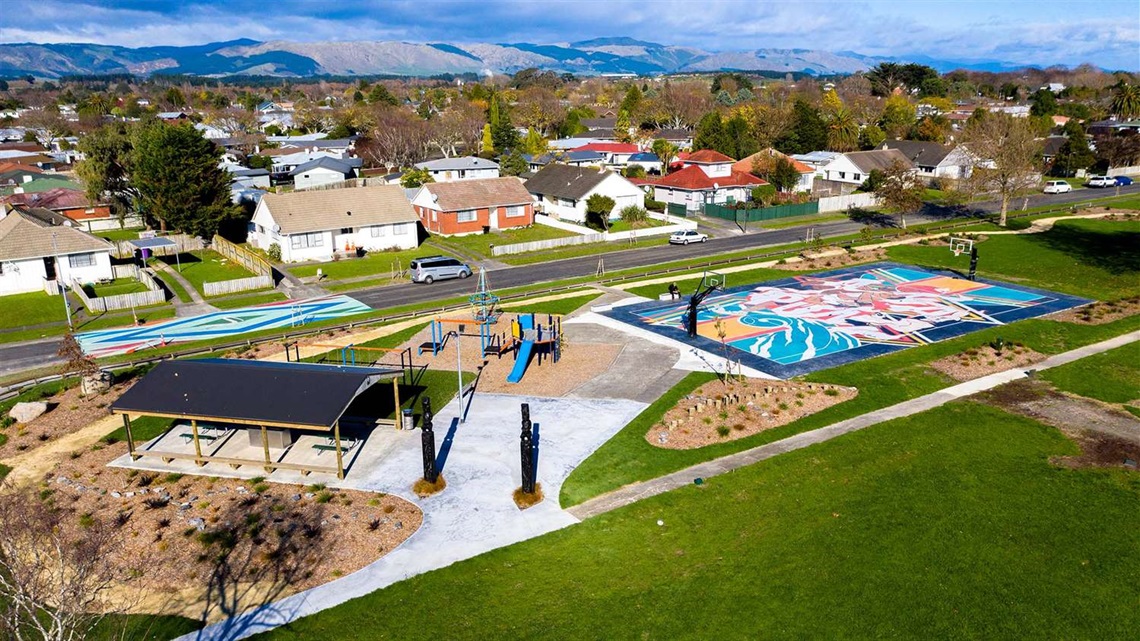 Photo shows a funky neighbourhood park with a basketball court painted with a colourful mural, a small playground and a pump track around the perimeter.