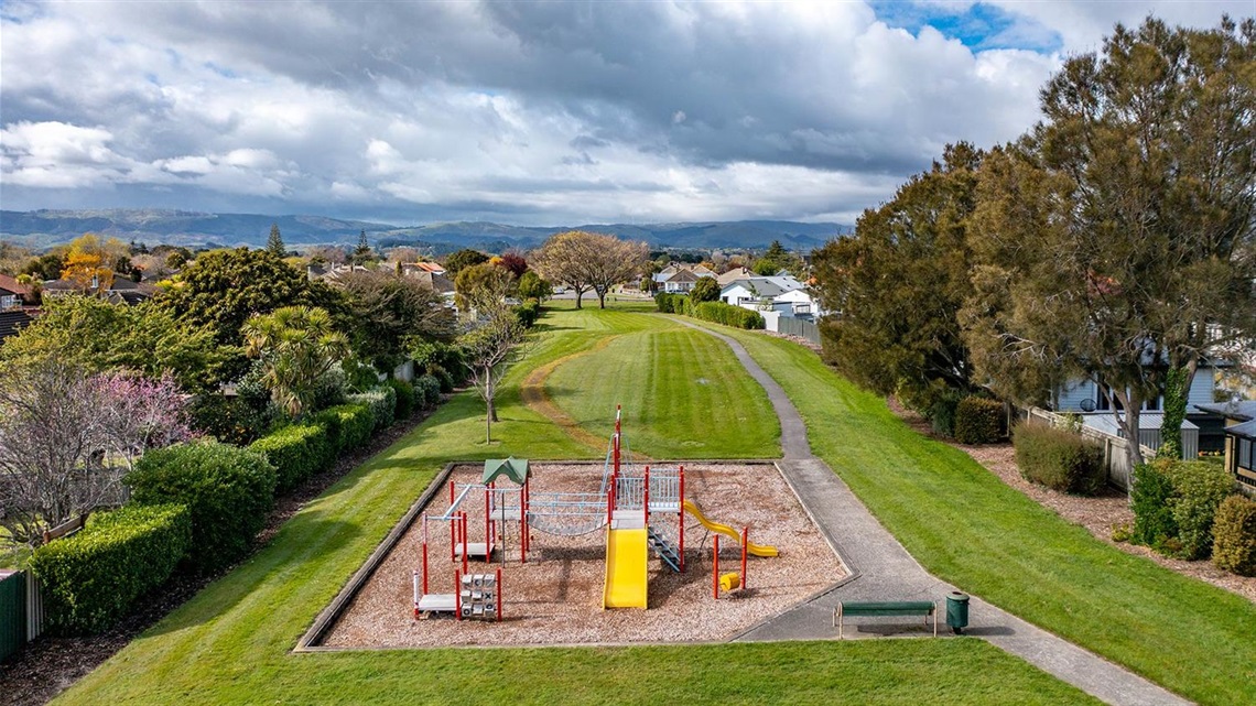 Picture shows a playground sitting in the front of a reserve, with a walkway running through the grassy land.