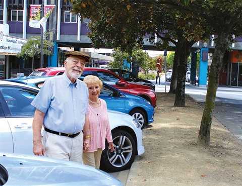 An older couple walking hand in hand in a PN city street lined with cars and trees.