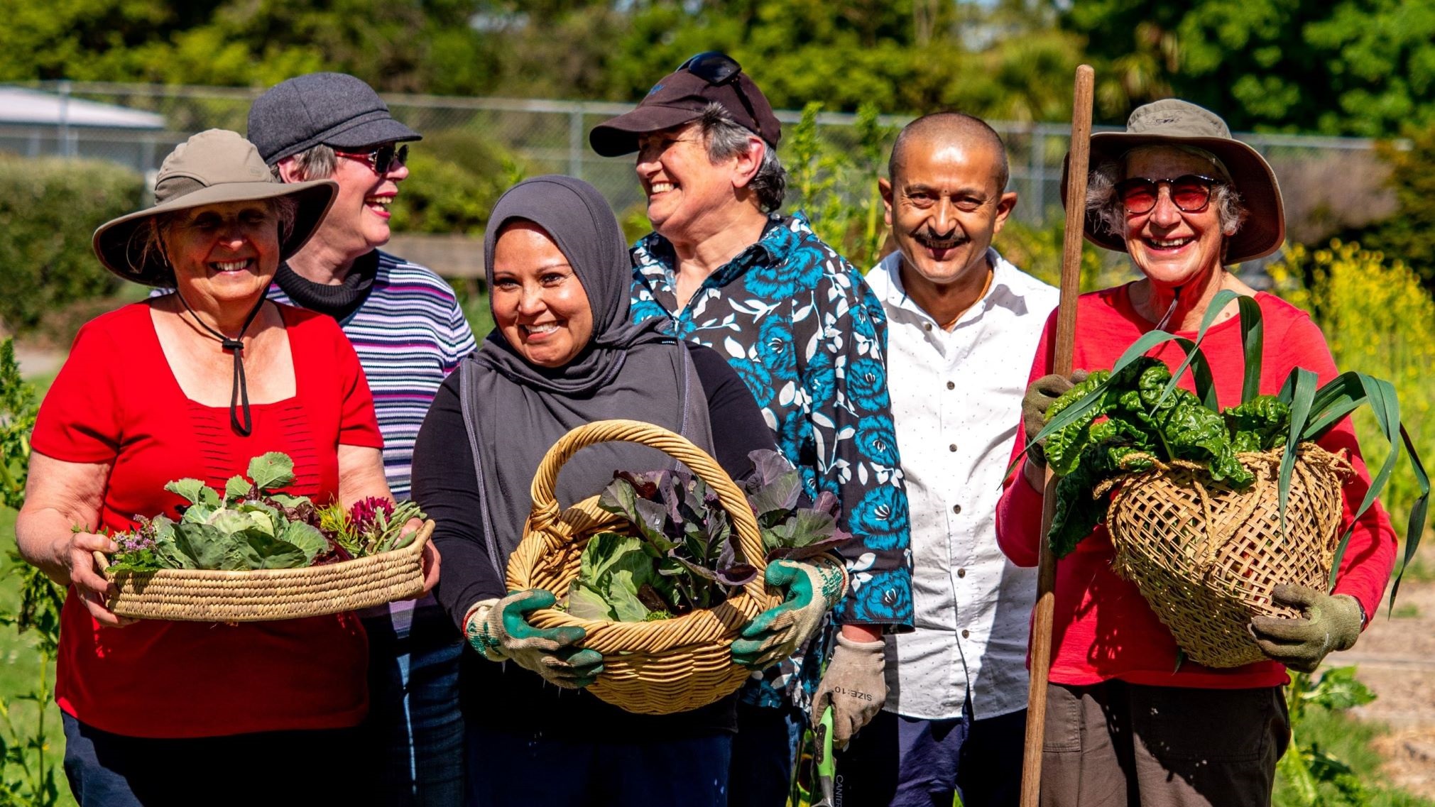Photo shows six smiling gardeners standing in a community garden holding baskets of fresh vegetables.