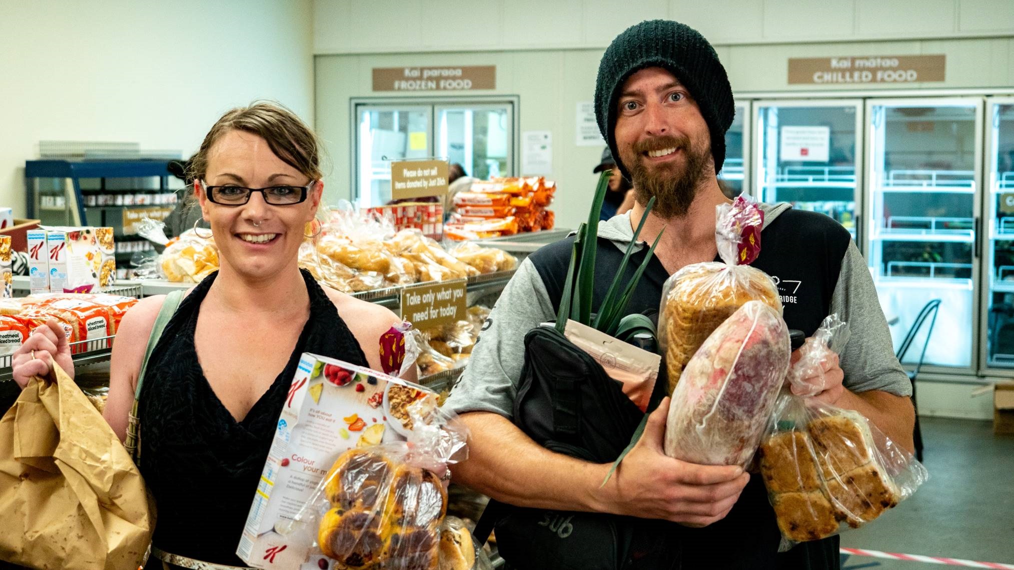 Photo shows Just Zilch patrons holding up bags of bread, meat and vegetables. There's plenty more on the shelves in the aisle behind them.