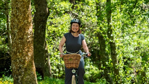 Smiling woman wearing flowing jumpsuit riding a utility bike with a wicker basket on the front through a bushy track.