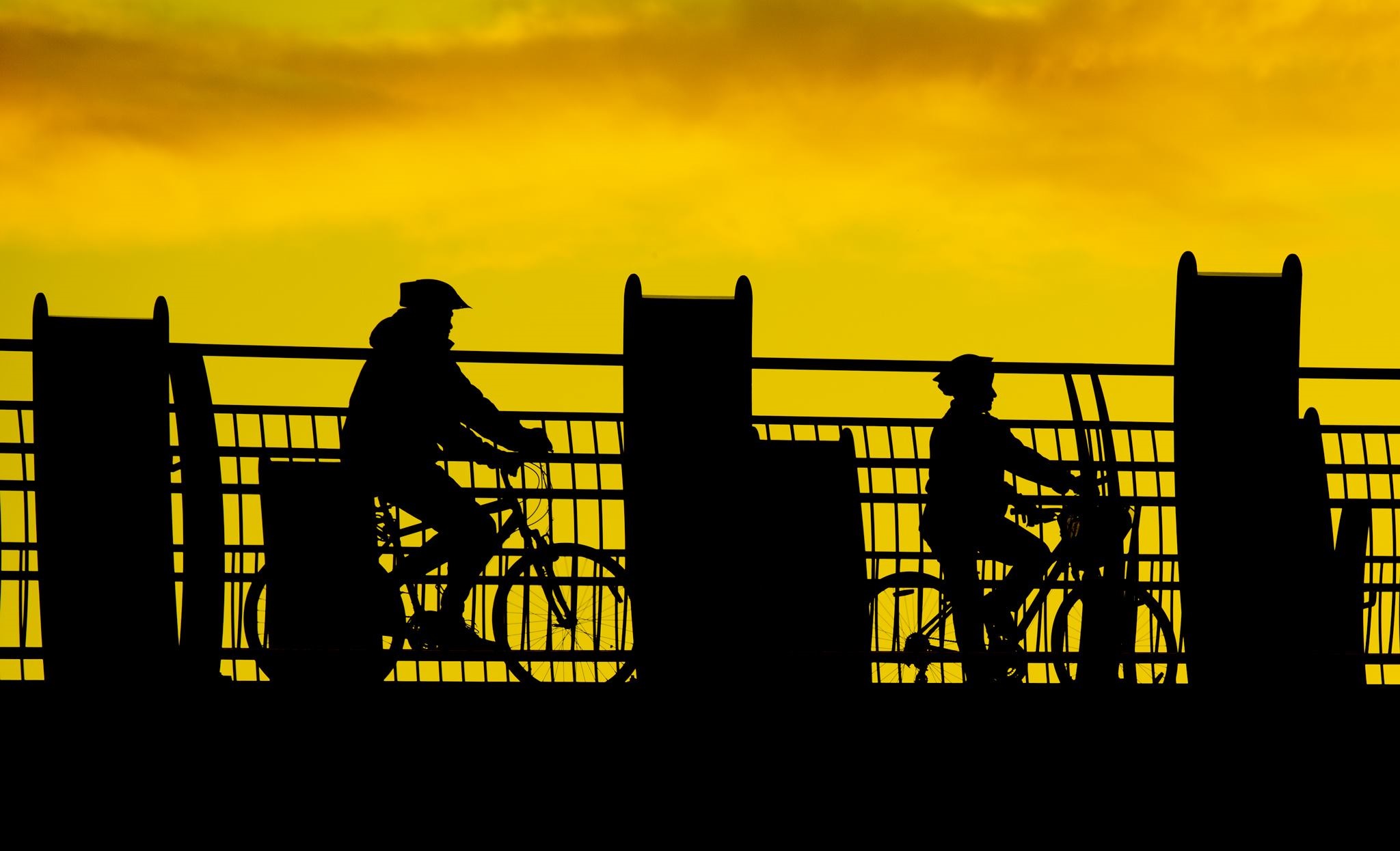 Photo shows two cyclists silhouetted against the skyline as they ride over a bridge.