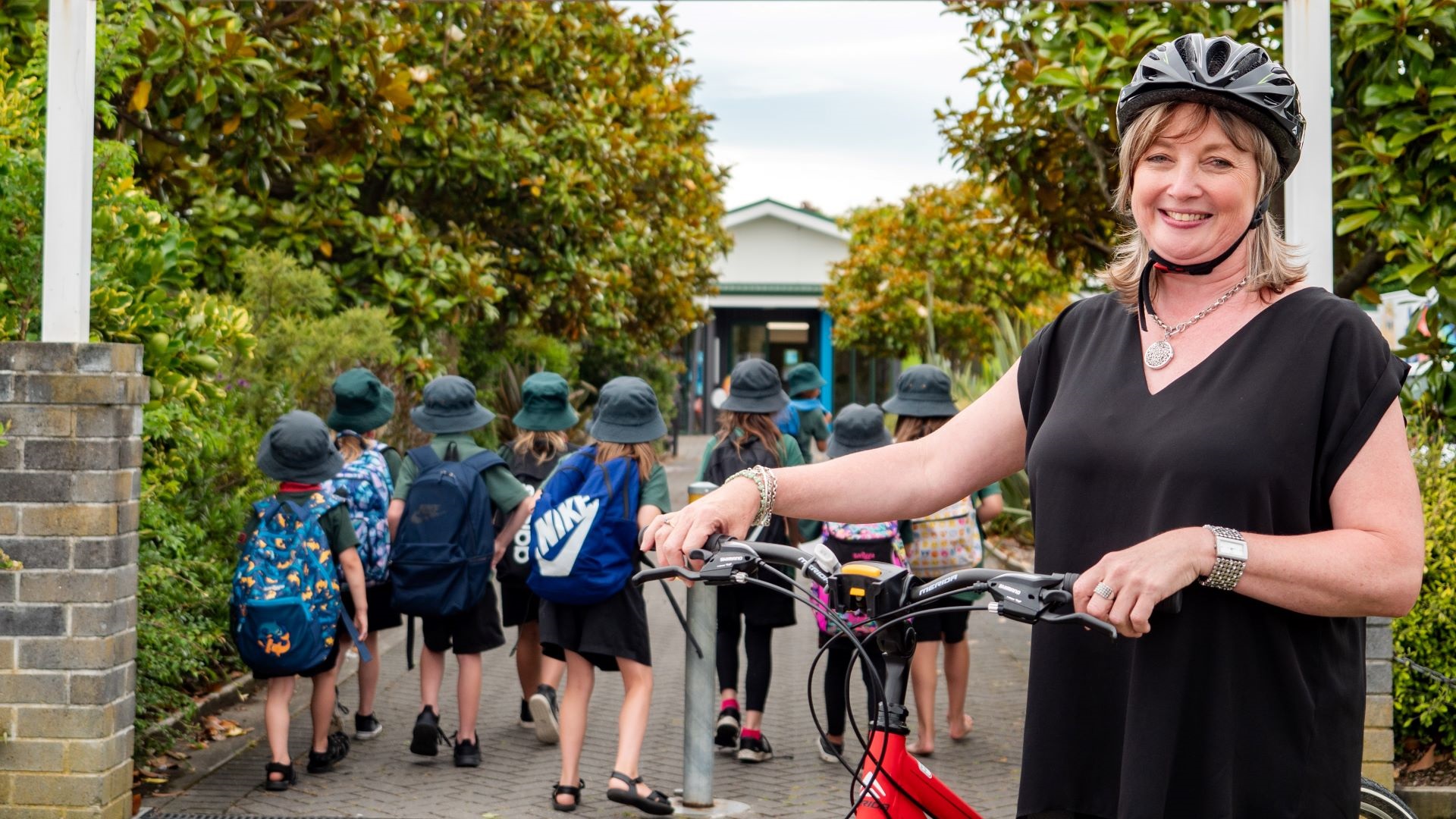 Photo shows smiling woman on bike at Cloverlea School gate with students in the background walking towards their classroom.