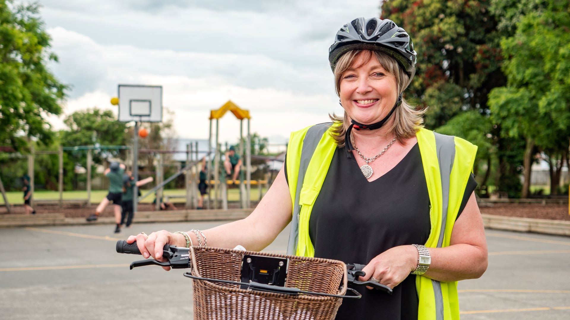 Photo shows smiling woman in helmet and hi vis holding bike with a wicker basket on the front.