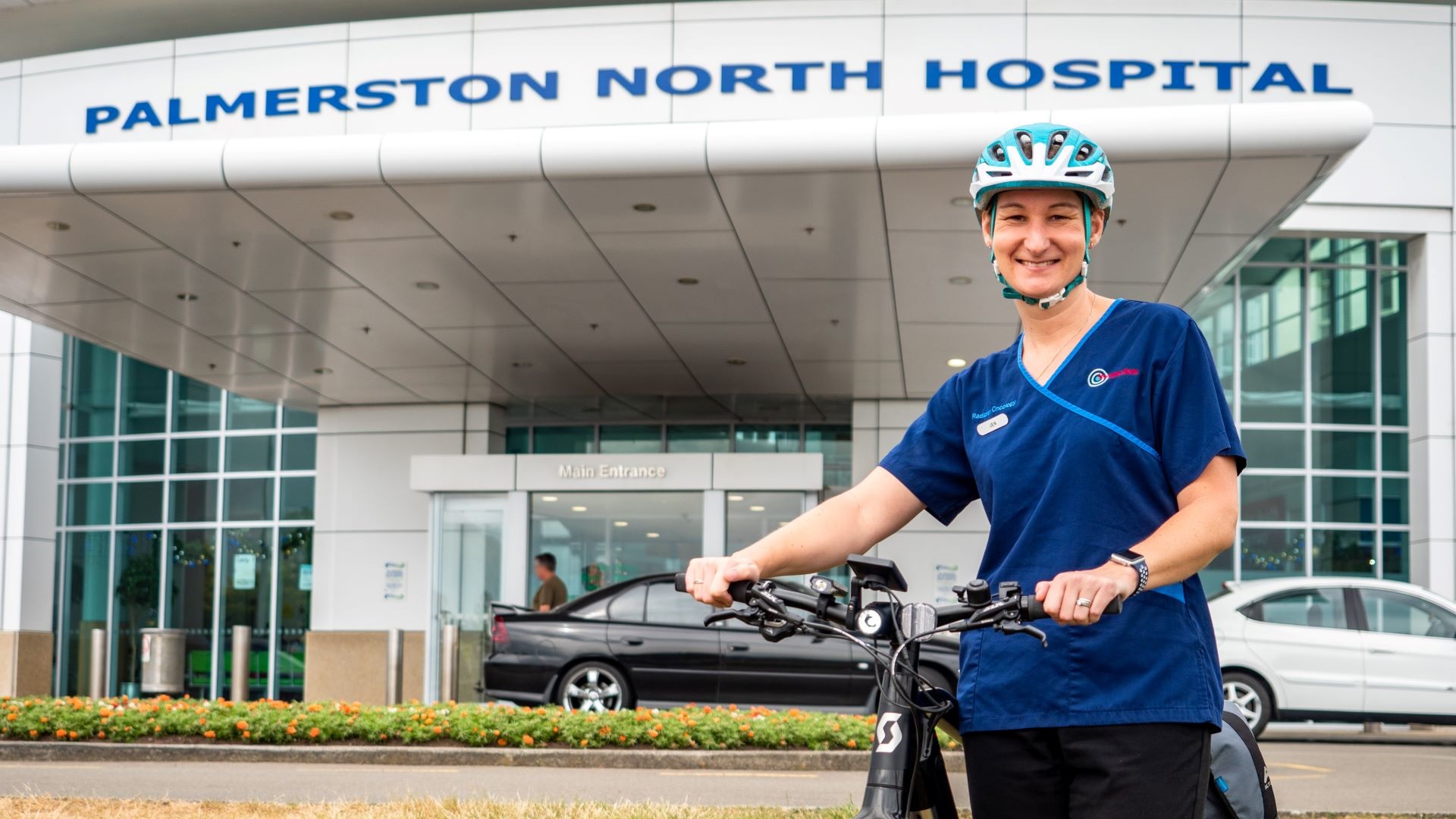 Photo shows smiling woman in hospital scrubs and bike helmet standing next to her bike outside the main entrance of Palmerston North Hospital.