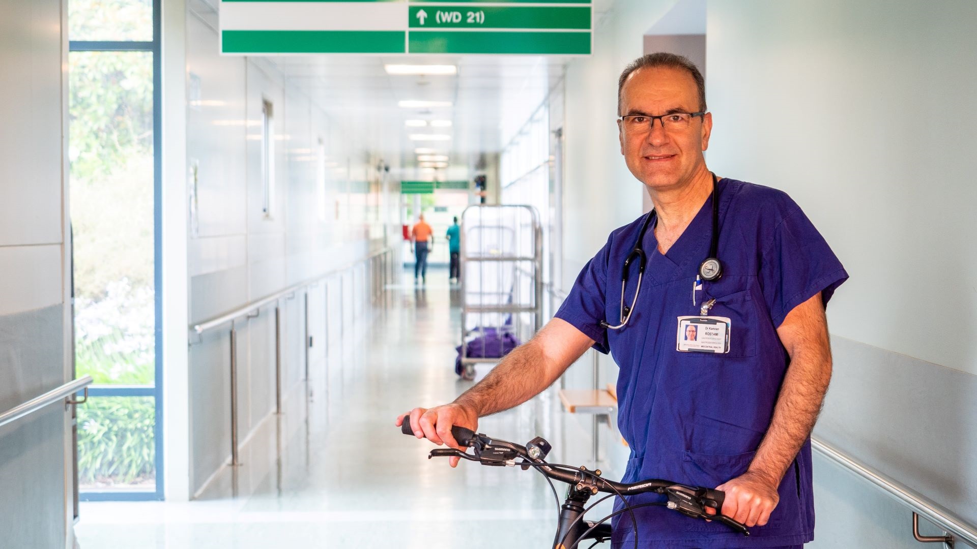 Photo shows man in hospital scrubs with stethoscope around his neck, posing with bike in a hospital corridor.