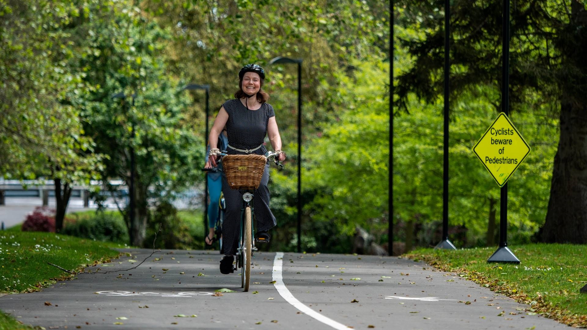 Photo shows woman in everyday clothes riding a commuter bike with a basket along a leafy off-road pathway.