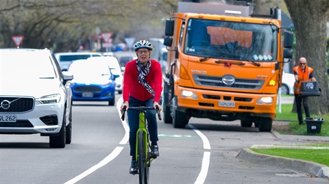 Woman cycling past a truck that's blocking part of the painted bike lane, while a heavy stream of cars passes on her right.