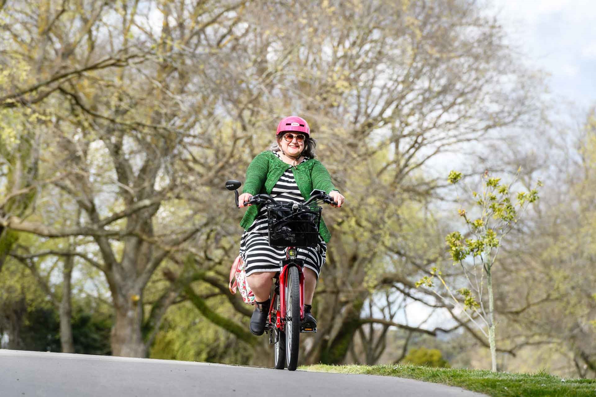 Photo shows smiling woman in a frock riding a utility bike with a flowery pannier and front basket along a tree-lined route.