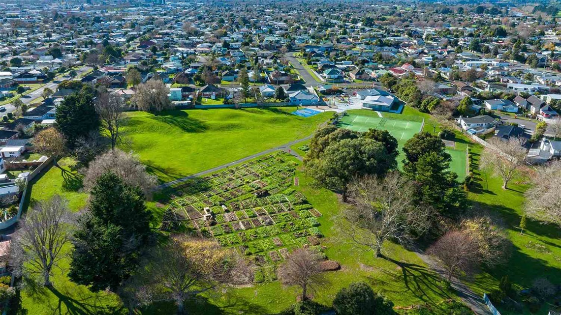 Photo shows aerial view of lawn tennis courts surrounded by shrubs with raised garden beds in the foreground.